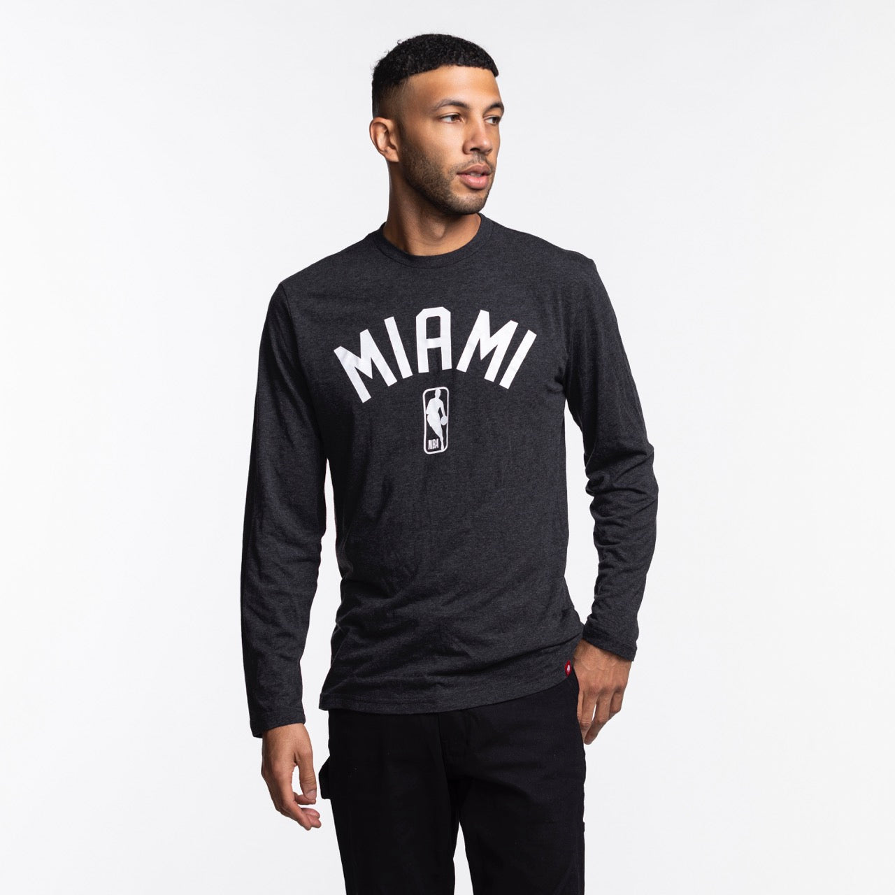 A man wears the Sportiqe Miami HEAT NBA Long Sleeve Tee—a black shirt by Sportiqe featuring MIAMI and an NBA logo. Made from a polyester-cotton-rayon blend, he stands against a white background, looking left with one hand in his pocket.