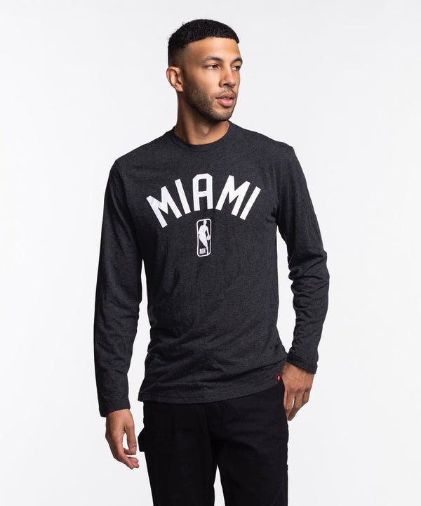A man wears the Sportiqe Miami HEAT NBA Long Sleeve Tee—a black shirt by Sportiqe featuring MIAMI and an NBA logo. Made from a polyester-cotton-rayon blend, he stands against a white background, looking left with one hand in his pocket.