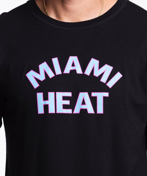 A man in a Sportiqe Miami HEAT Original Vice Nights Long Sleeve Tee stands against a white background. The black cotton shirt has pink and white MIAMI HEAT text. He has short dark hair, light facial hair, and wears blue jeans.