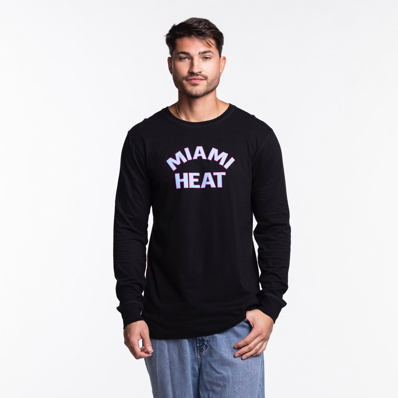 A man in a Sportiqe Miami HEAT Original Vice Nights Long Sleeve Tee stands against a white background. The black cotton shirt has pink and white MIAMI HEAT text. He has short dark hair, light facial hair, and wears blue jeans.