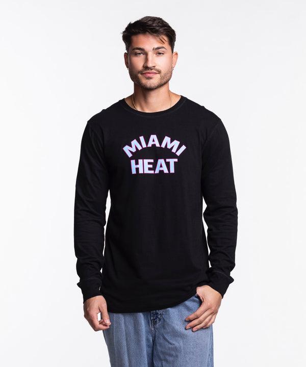 A man in a Sportiqe Miami HEAT Original Vice Nights Long Sleeve Tee stands against a white background. The black cotton shirt has pink and white MIAMI HEAT text. He has short dark hair, light facial hair, and wears blue jeans.