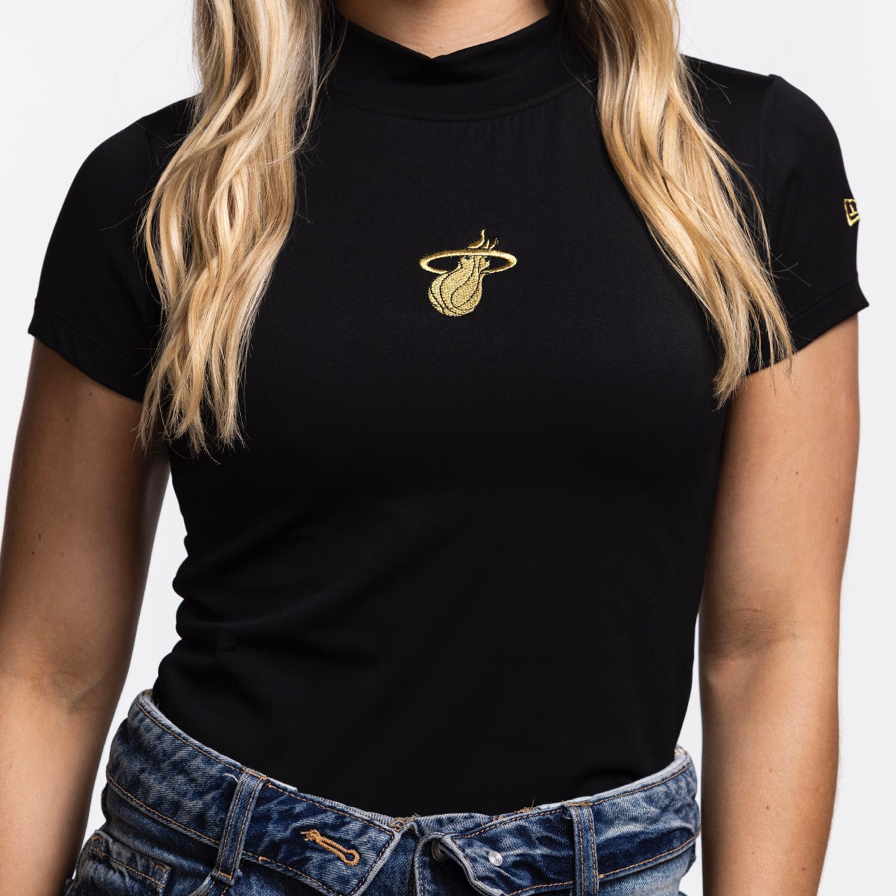 A woman wears the New Era Miami HEAT Women's Mock Neck Tee, featuring a fitted black design with a gold logo, paired with blue jeans. Her loose long blonde hair partially covers her shoulders.