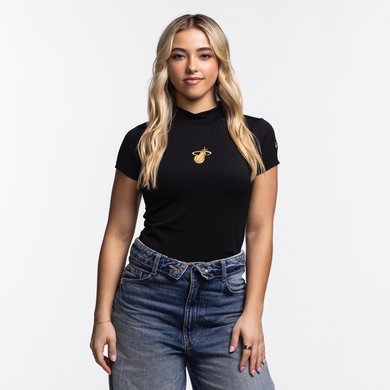 A woman with long blonde hair wears the New Era Miami HEAT Women's Mock Neck Tee, a fitted black t-shirt with a small yellow logo, paired with blue jeans. She faces the camera against a plain white background. Brand: New Era.