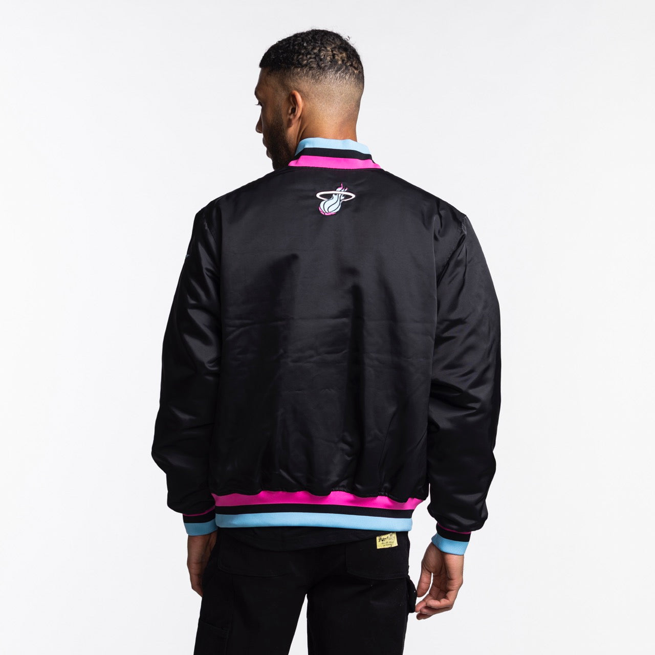 A man stands with his back turned, wearing the Nike Miami HEAT Original Vice Nights Bomber Jacket—an insulated black polyester design with pink and blue trim and a Miami Heat logo—against a plain white background.