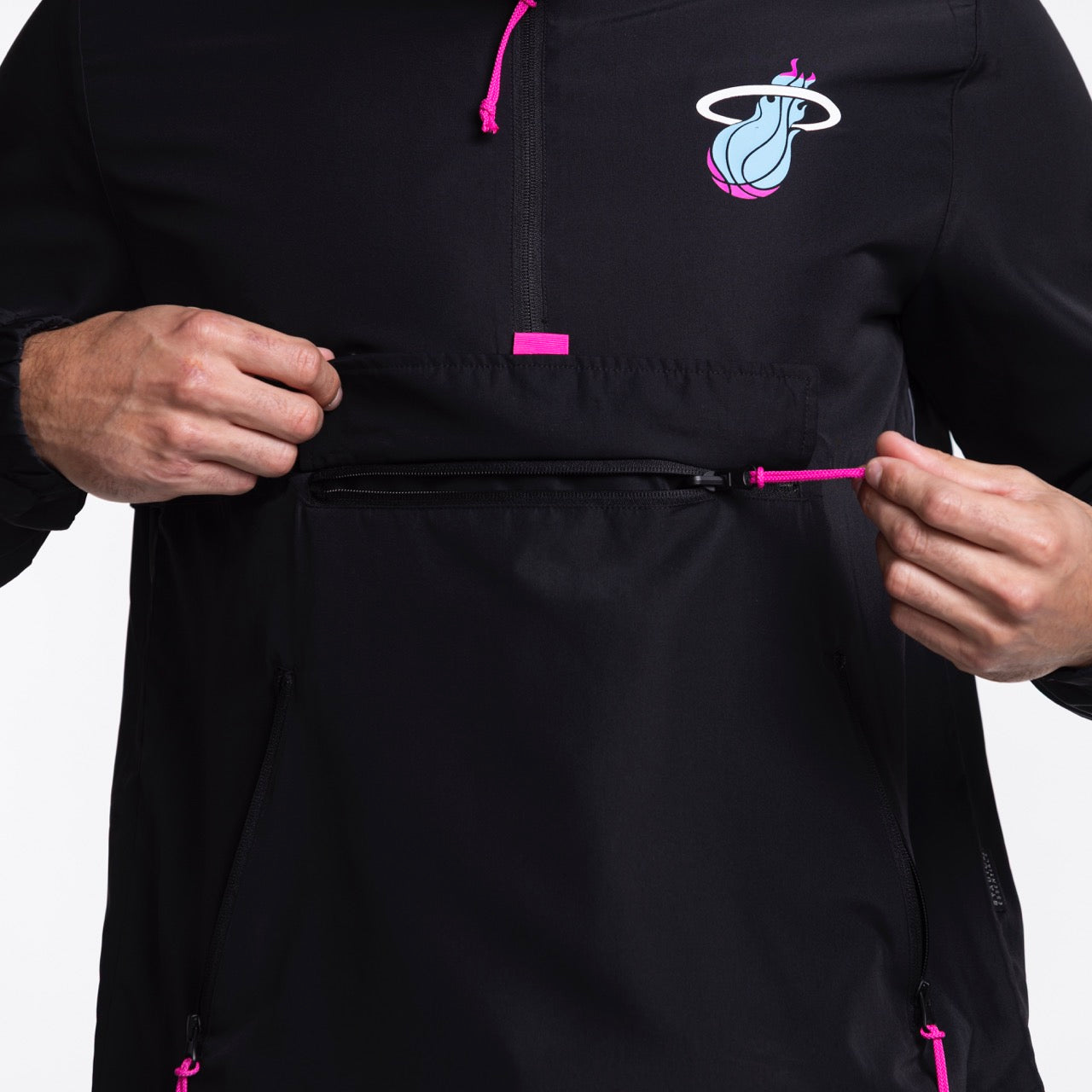 Someone unzips the front pocket of the black Stadium Essentials Miami HEAT Original Vice Nights 1/4 Zip Jacket, featuring pink zippers and a teal, pink, and white team logo—easy to spot clean and pairs great with a polyester hat.