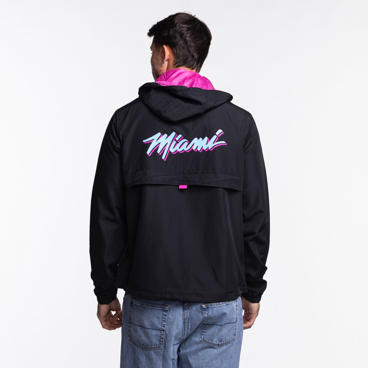 A person wearing a Stadium Essentials Miami HEAT Original Vice Nights 1/4 Zip Jacket with “Miami” in a colorful retro font on the back, blue jeans, and a polyester hat stands facing away against a plain white background.