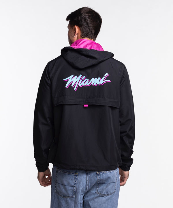 A man wears the Stadium Essentials Miami HEAT Original Vice Nights 1/4 Zip Jacket—black with pink accents and a Miami Heat logo—paired with a polyester hat, standing against a plain white background. One size fits most; spot clean only.