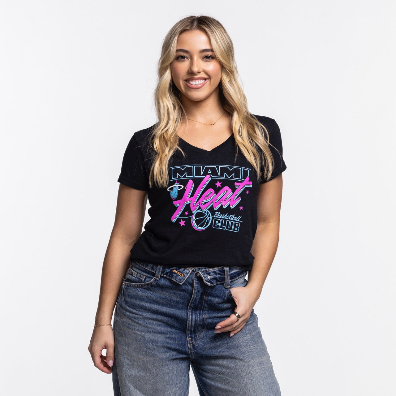 A smiling woman with long blonde hair wears the Miami HEAT Original Vice Nights Basketball Club Women's Tee by Item Of The Game, made from 100% cotton, with blue jeans, standing against a plain white background.