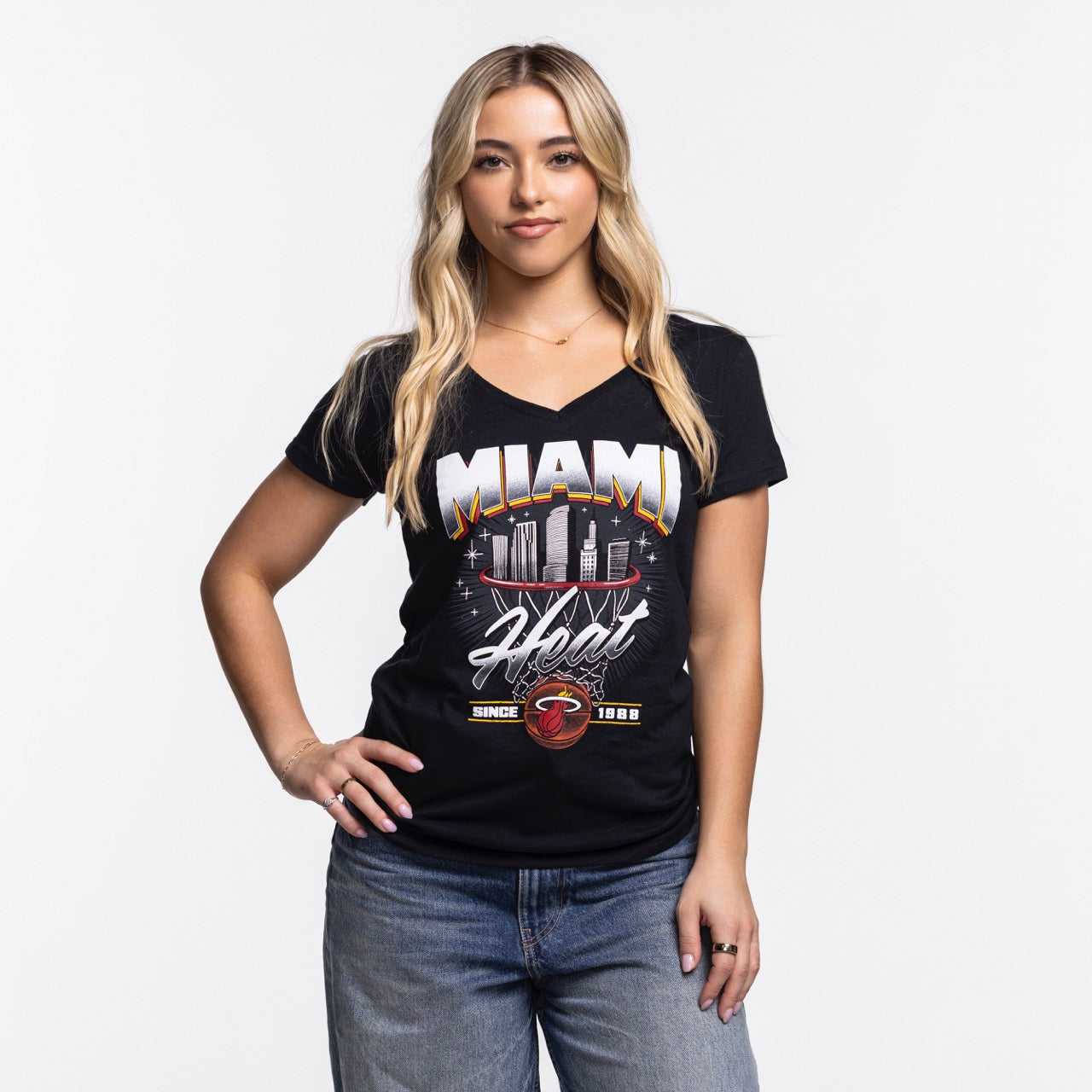 A woman with long blonde hair, wearing the Item Of The Game Miami HEAT Women's Retro Net Tee and blue jeans, stands against a plain white background with one hand on her hip and a relaxed expression.