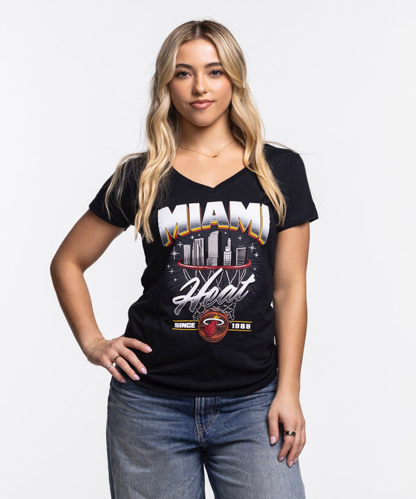 A woman with long blonde hair, wearing the Item Of The Game Miami HEAT Women's Retro Net Tee and blue jeans, stands against a plain white background with one hand on her hip and a relaxed expression.