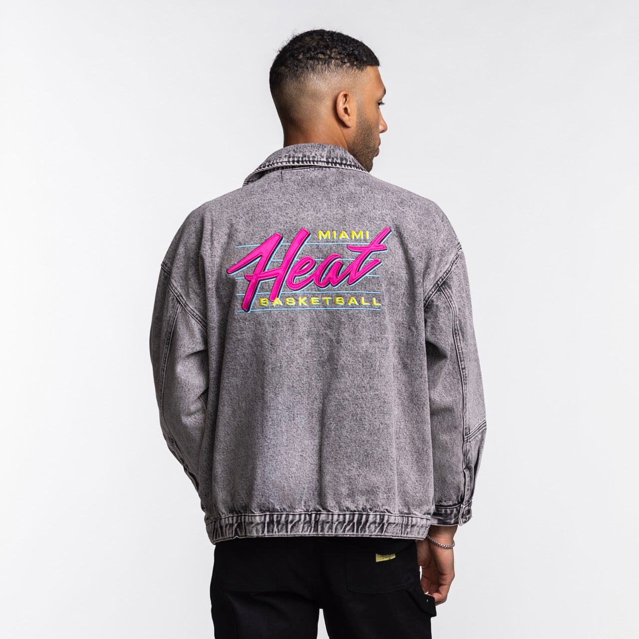 A man faces away, wearing the Court Culture X The Wild Collective Original Vice Nights Denim Patch Unisex Jacket in light gray cotton. It features a pink and yellow Miami Heat logo on the back. Dry clean only. Brand: Court Culture.