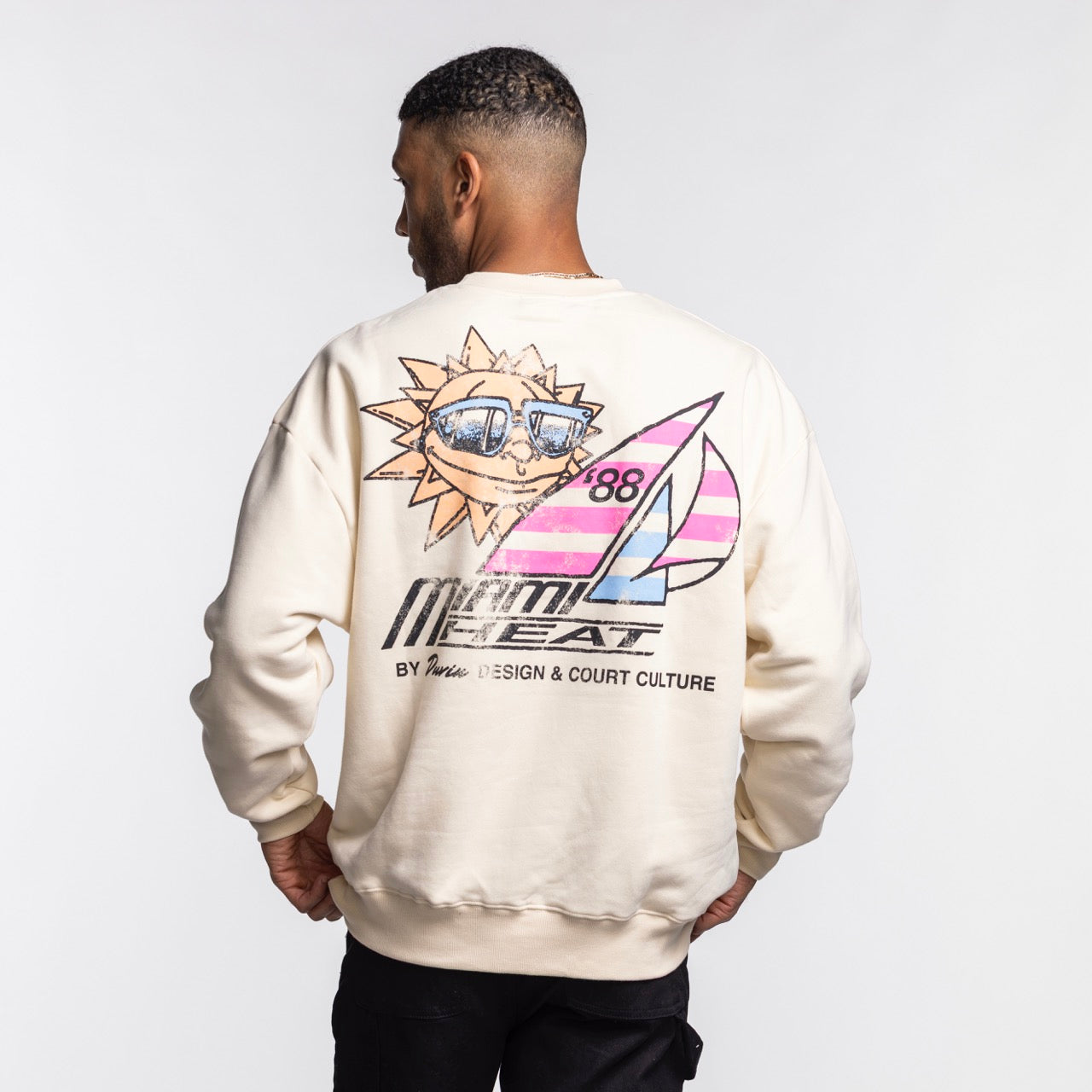 A man faces away in the Court Culture x Duvin Sun Crew Sweater by Court Culture, part of the NBA Official Collection, with a colorful smiling sun, sailboat, and bold “Miami Heat” graphic on the back.