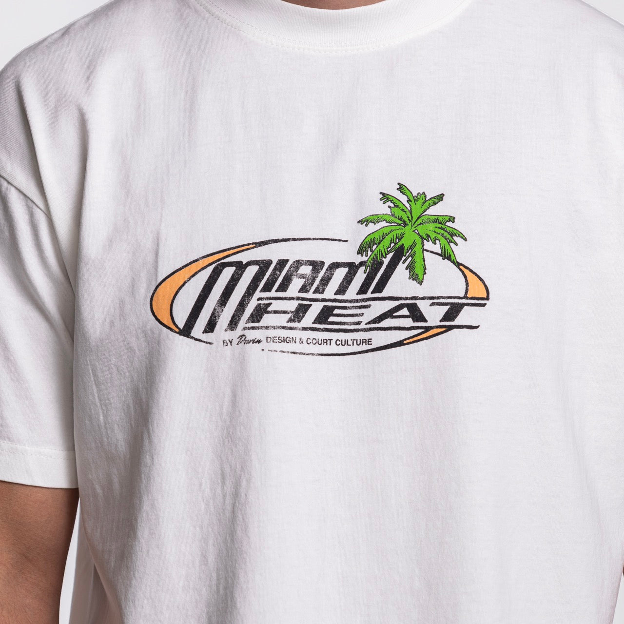 Someone wearing the white Court Culture x Duvin Sun Tee by Court Culture, featuring “Miami Heat” on the front with a green palm tree and orange oval graphic from the NBA Official Collection in collaboration with Duvin Design Co.