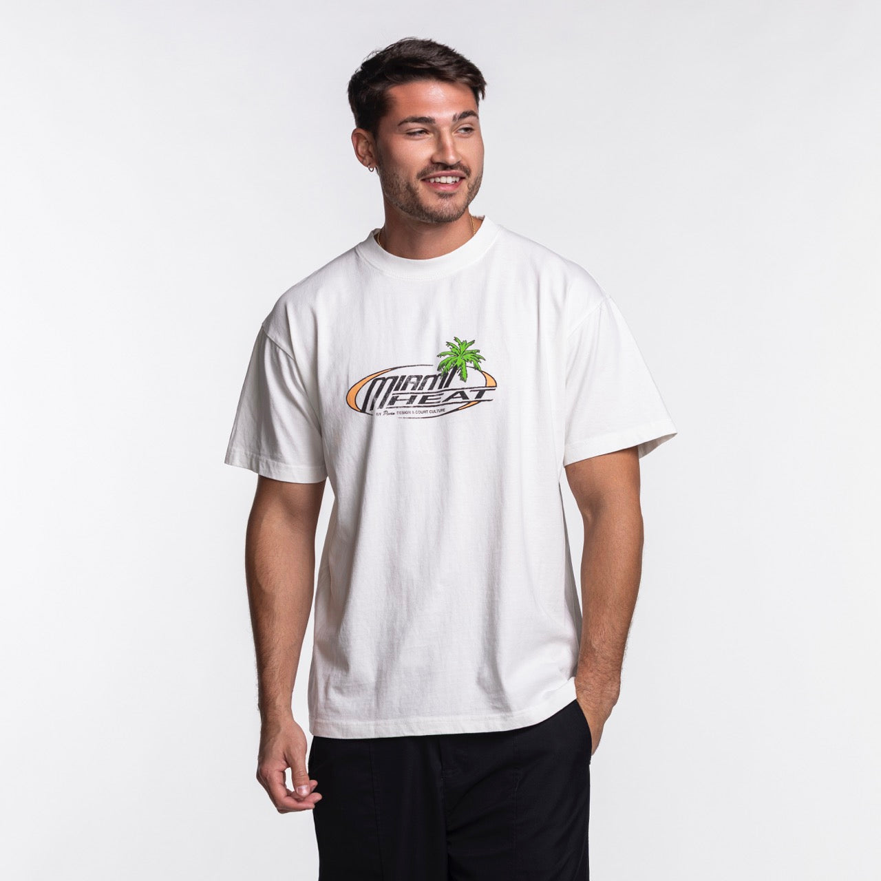 A man stands against a plain white background, wearing the Court Culture x Duvin Sun Tee from Court Culture with a Miami Heat logo and palm tree graphic, paired with black pants. He looks to the side and smiles.