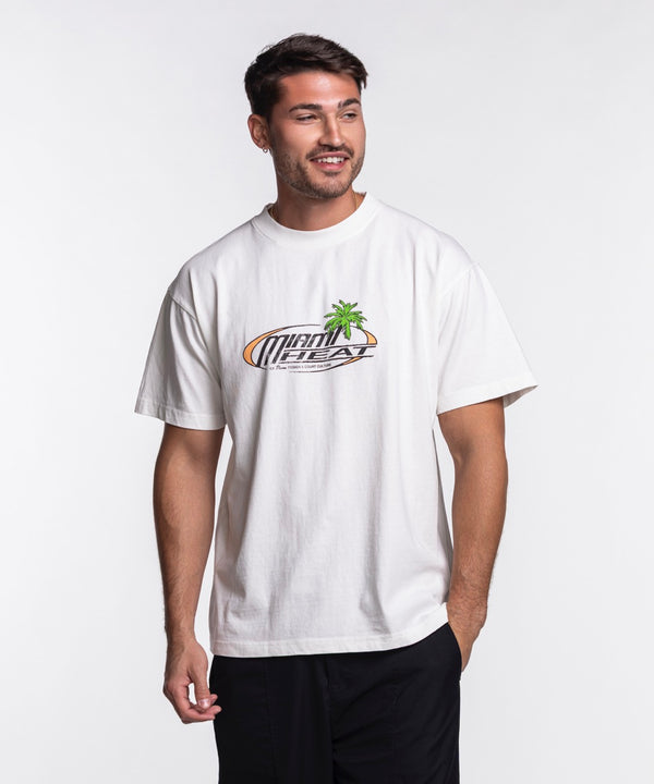 A man stands against a plain white background, wearing the Court Culture x Duvin Sun Tee from Court Culture with a Miami Heat logo and palm tree graphic, paired with black pants. He looks to the side and smiles.