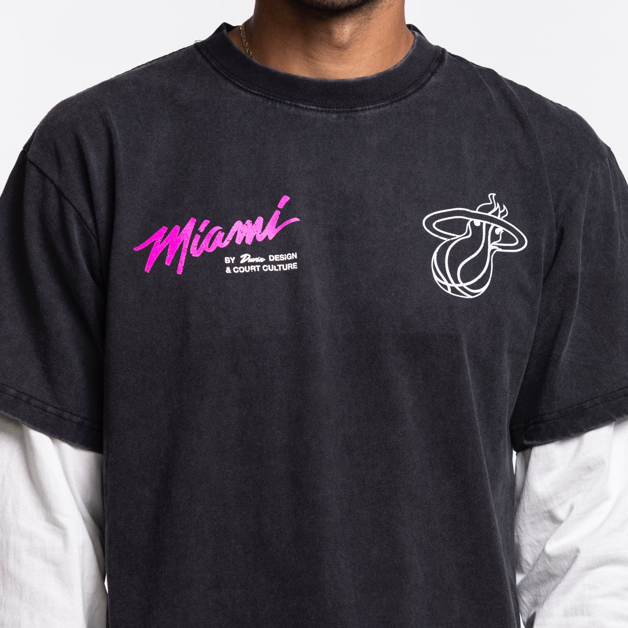 A person wearing a dark gray Court Culture x Duvin Original Vice Long Sleeve Tee, part of the Court Culture collection, featuring Miami in pink script and a white flaming basketball logo, styled over a white long-sleeve shirt.