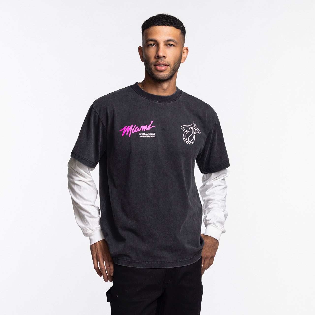 A man stands against a white background wearing a black Court Culture x Duvin Original Vice Long Sleeve Tee by Court Culture, featuring pink Vice Nights lettering, layered over a white long-sleeve shirt with black pants.