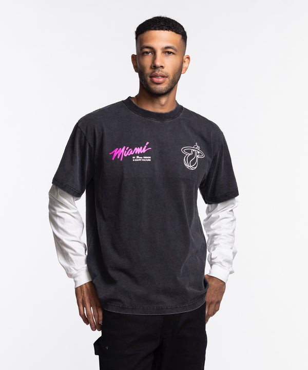 A man stands against a white background wearing a black Court Culture x Duvin Original Vice Long Sleeve Tee by Court Culture, featuring pink Vice Nights lettering, layered over a white long-sleeve shirt with black pants.