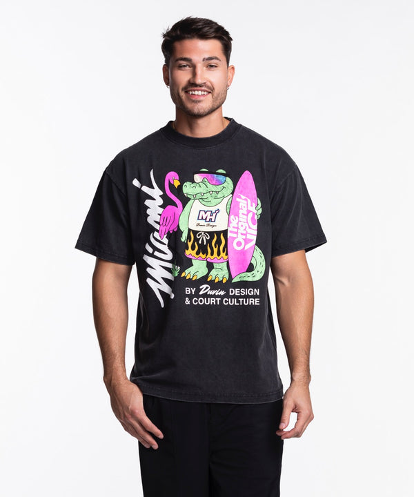 A man stands against a white background wearing the Court Culture x Duvin Flame Surf Tee by Court Culture, a black heavyweight shirt featuring colorful graphics of a cartoon crocodile, flamingo, surfboard, and 