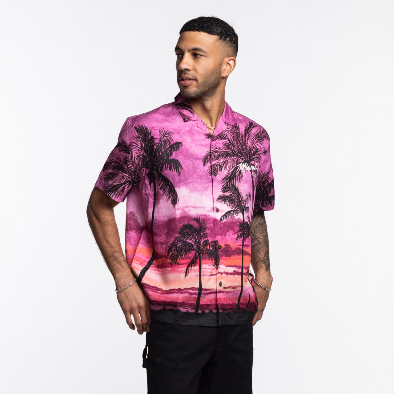 A man wears the Court Culture x Duvin Sunset Button-Up, featuring a vibrant pink and purple sunset with palm tree design, standing against a white background and looking left with one hand in his pocket.