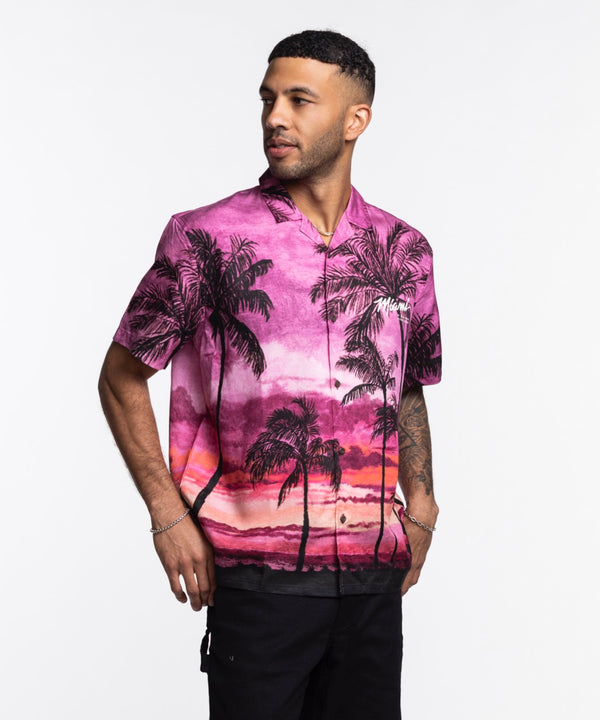 A man wears the Court Culture x Duvin Sunset Button-Up, featuring a vibrant pink and purple sunset with palm tree design, standing against a white background and looking left with one hand in his pocket.