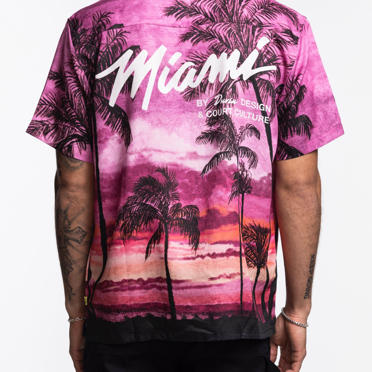 A person wears the Court Culture x Duvin Sunset Button-Up, a pink and purple shirt featuring black palm trees and 