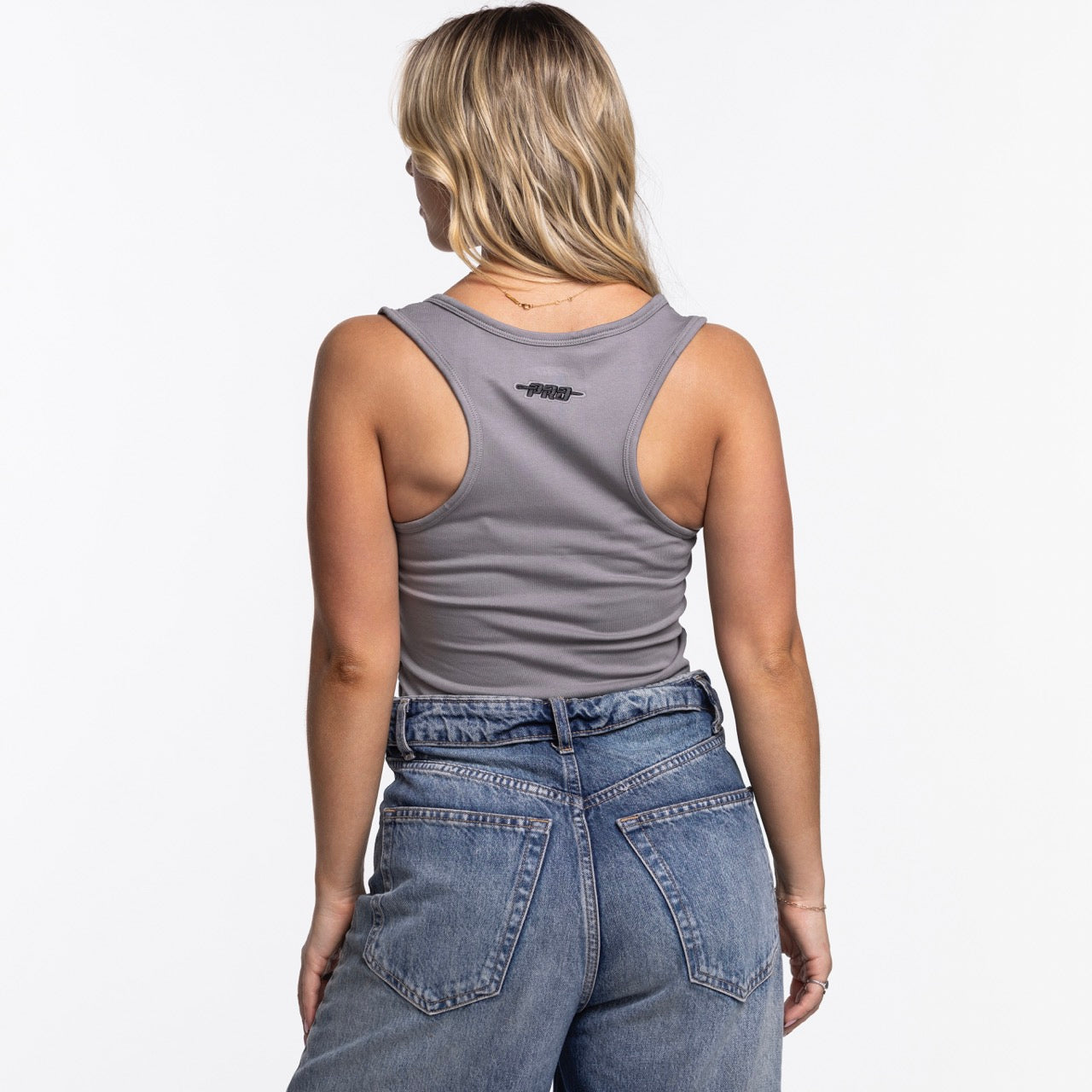 A woman with blonde hair stands with her back to the camera against a plain white background, wearing the Pro Standard Miami HEAT Original Vice Nights Women's Tank by Pro Standard and high-waisted blue jeans.