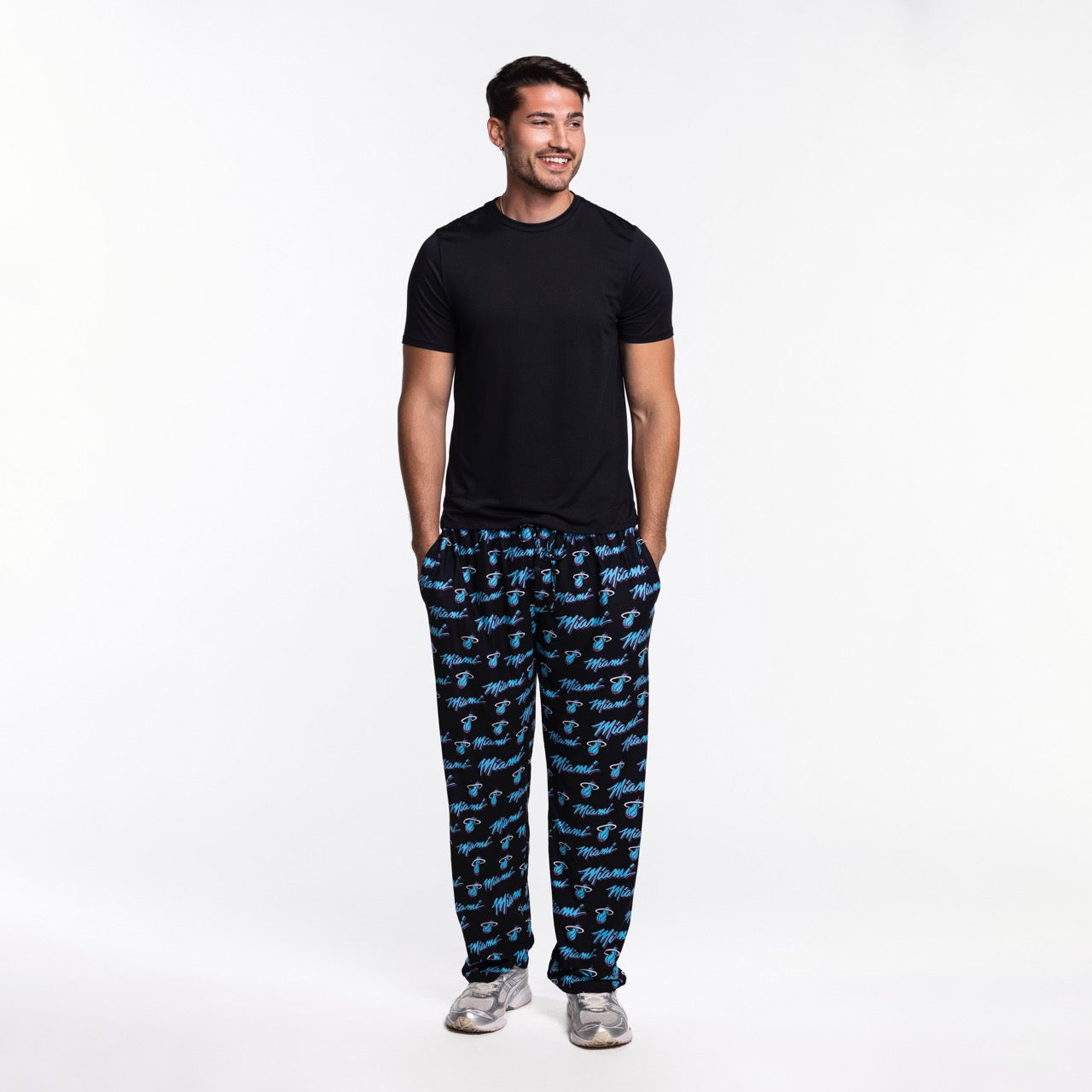 A man stands smiling with his hands in the pockets of his Concepts Sports Miami HEAT Original Vice Nights Logo Pants, paired with a black t-shirt and sneakers against a plain white background.