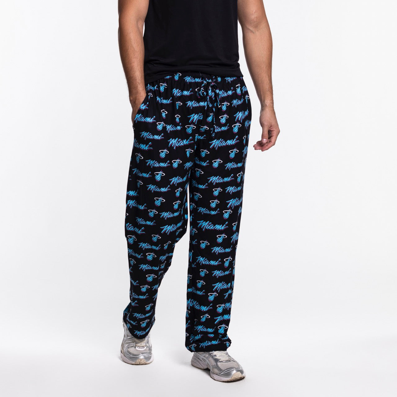 A person wears Concepts Sports Miami HEAT Original Vice Nights Logo Pants—relaxed fit, black with blue Heat logos, made from soft polyester—paired with a black t-shirt and silver sneakers, standing against a plain white background.