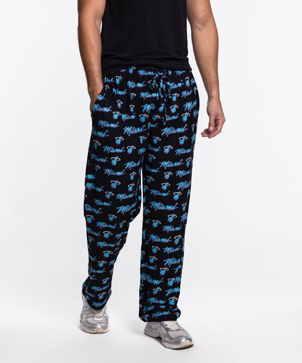 A person wears Concepts Sports Miami HEAT Original Vice Nights Logo Pants—relaxed fit, black with blue Heat logos, made from soft polyester—paired with a black t-shirt and silver sneakers, standing against a plain white background.