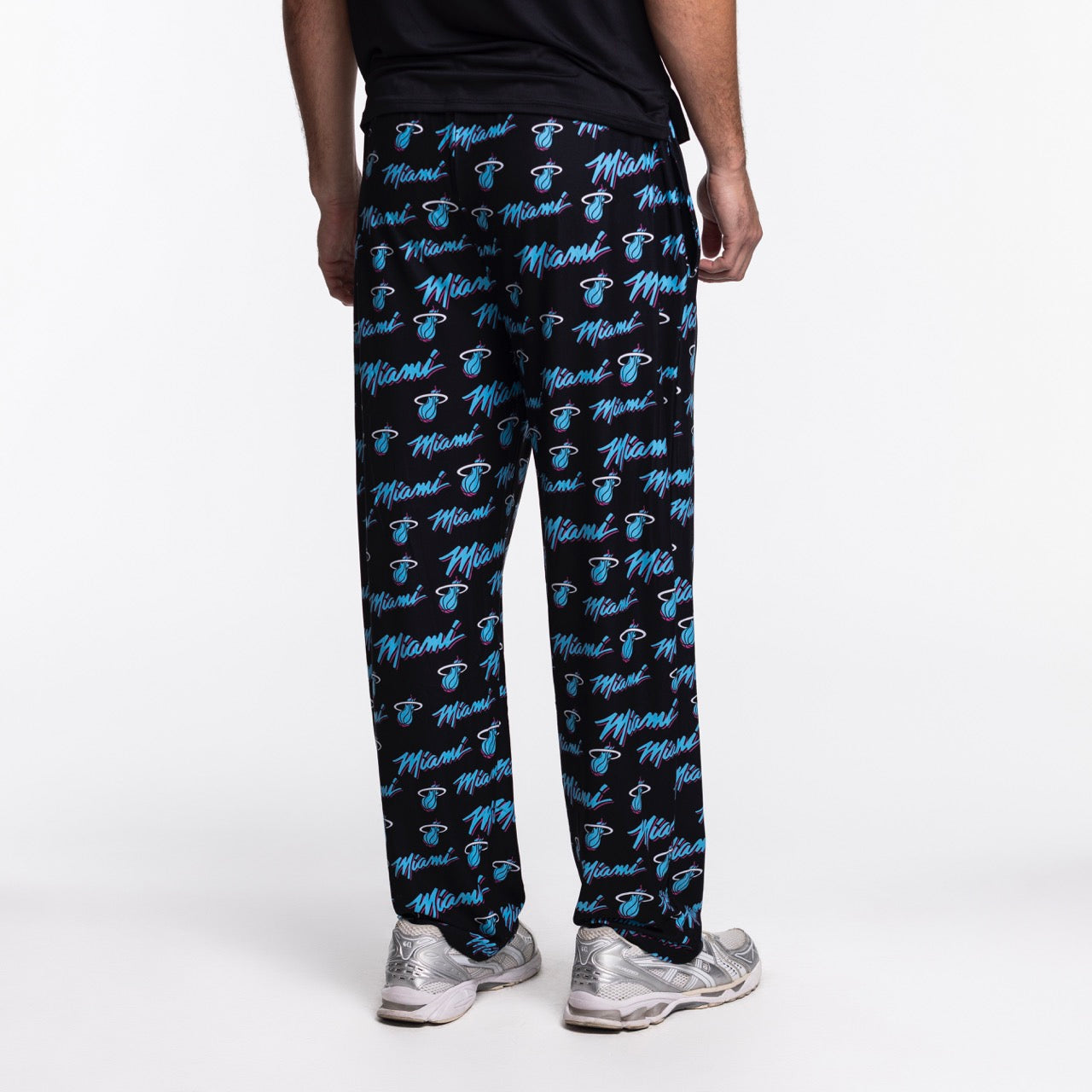 Someone stands with their back to the camera wearing Concepts Sports Miami HEAT Original Vice Nights Logo Pants—black pajama pants with blue Miami text and basketball logos, made from a relaxed fit polyester-spandex blend—paired with a black shirt and silver sneakers.