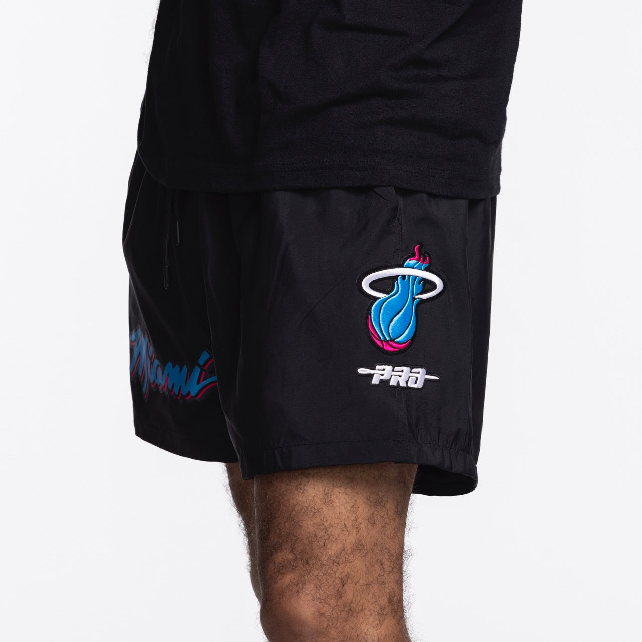A person is shown from the waist down wearing Pro Standard Miami HEAT Original Vice Nights Woven Shorts in black, featuring the flaming basketball logo and 