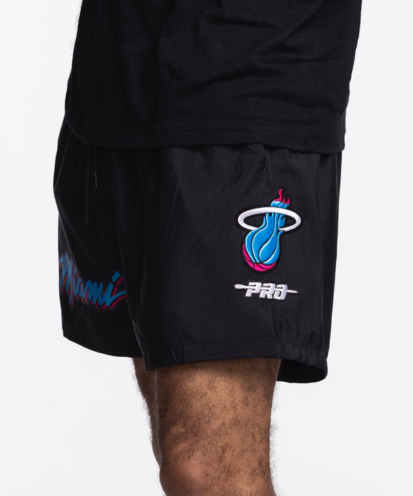 A person is seen from the torso down wearing Pro Standard Miami HEAT Original Vice Nights Woven Shorts, featuring colorful 'Miami' lettering on the left leg and paired with a black shirt; both pieces have a standard fit.