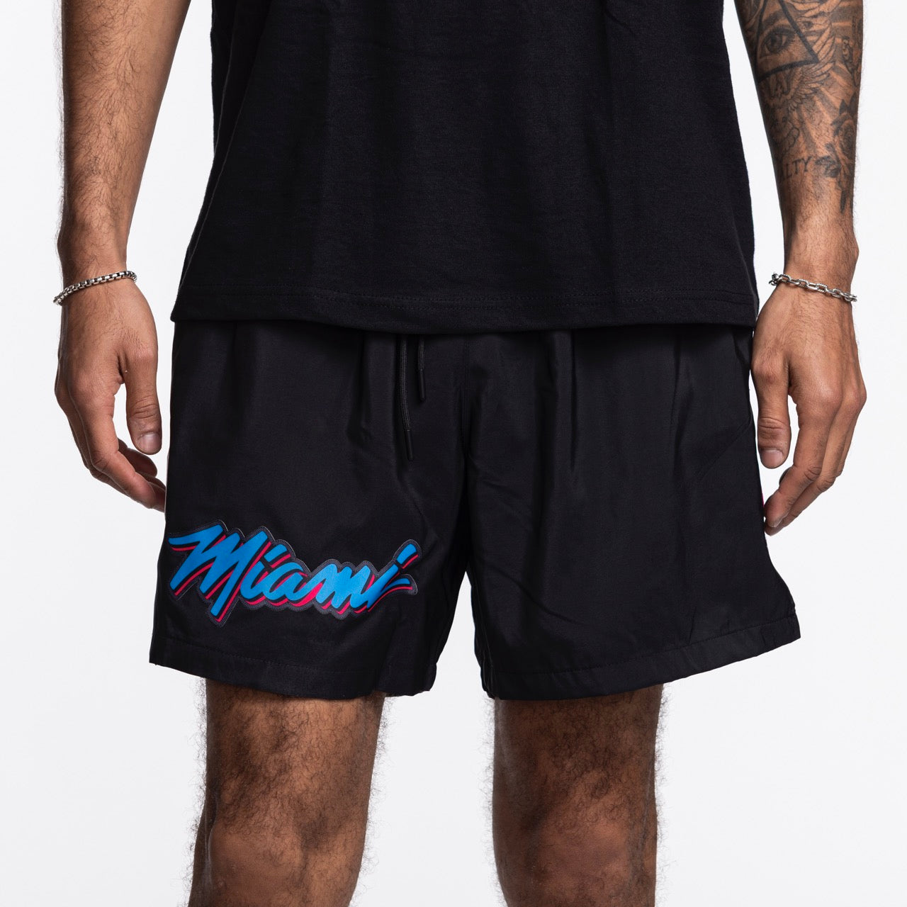 A person is seen from the torso down wearing Pro Standard Miami HEAT Original Vice Nights Woven Shorts, featuring colorful 'Miami' lettering on the left leg and paired with a black shirt; both pieces have a standard fit.