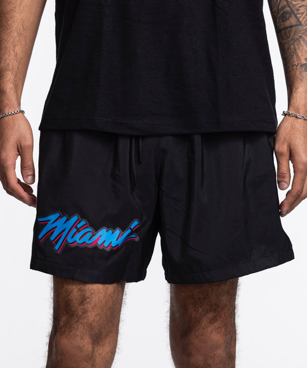 A person is seen from the torso down wearing Pro Standard Miami HEAT Original Vice Nights Woven Shorts, featuring colorful 'Miami' lettering on the left leg and paired with a black shirt; both pieces have a standard fit.