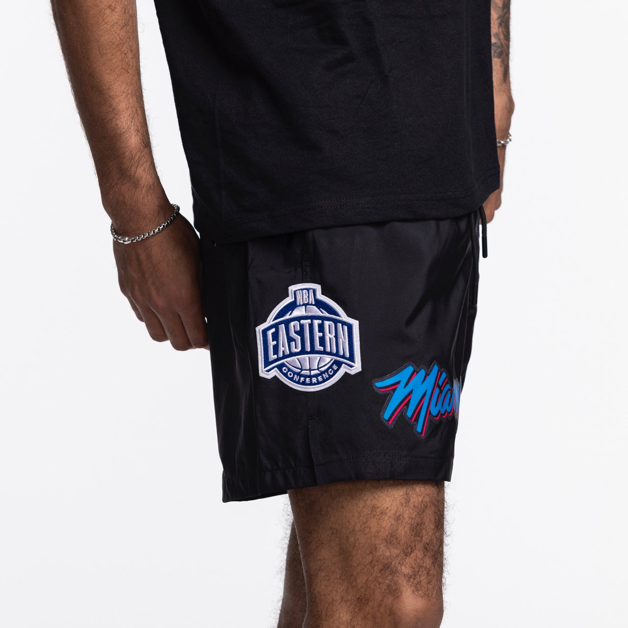 A person in Pro Standard Miami HEAT Original Vice Nights Woven Shorts featuring the NBA Eastern Conference patch and team logo, standing sideways in a black shirt against a plain white background.
