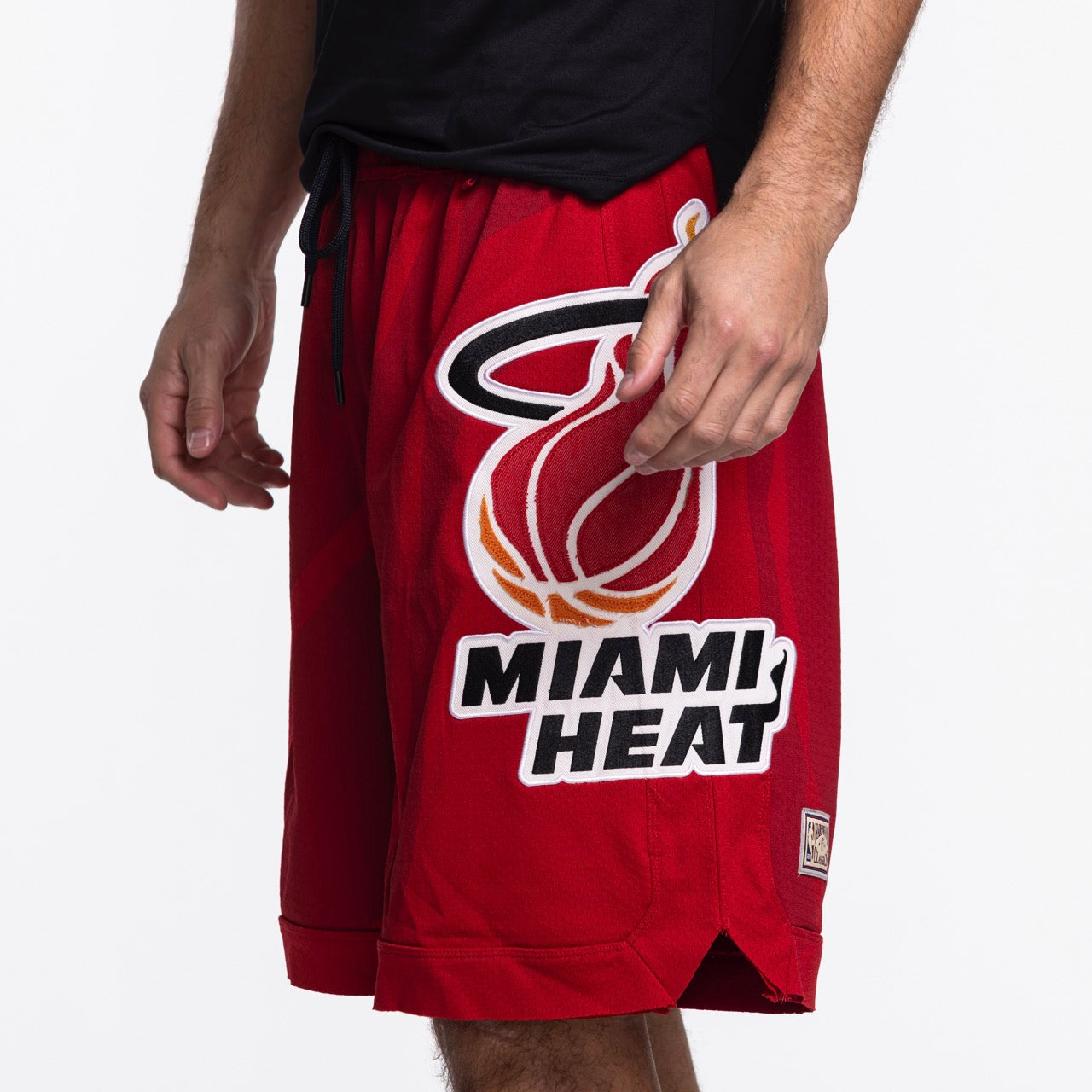 Someone is seen in Mitchell & Ness x Purple Brand Miami HEAT Mesh Shorts, featuring the team logo and text on the left leg. The person’s upper body, partly shown, wears a black shirt.
