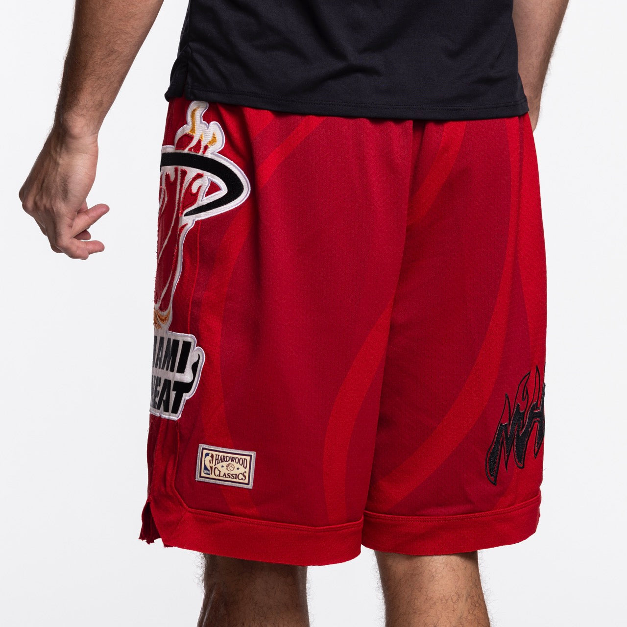Someone wearing Mitchell & Ness x Purple Brand Miami HEAT Mesh Shorts, with team logos and a Hardwood Classics patch, stands facing away to highlight the back and side of these iconic red premium sport mesh shorts.