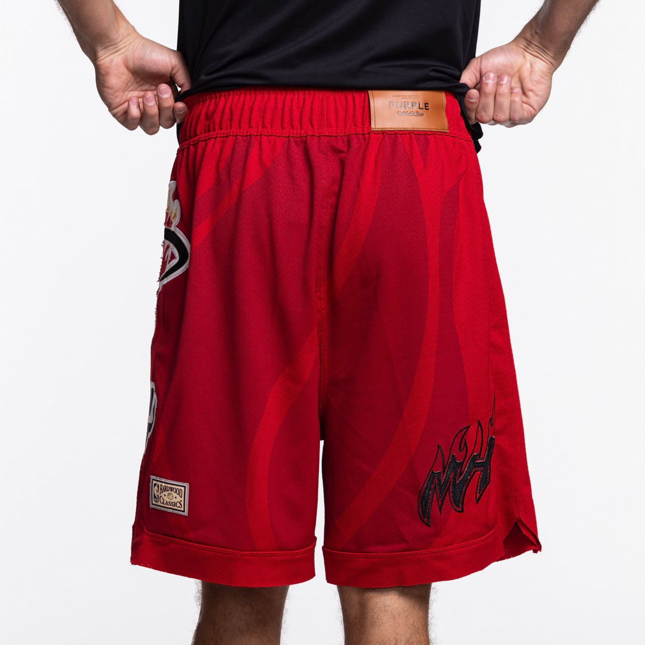 A person wears Mitchell & Ness x Purple Brand Miami HEAT Mesh Shorts—bright red sport mesh with a leather waistband patch, sports logos, and a flame graphic—standing against a plain white background.