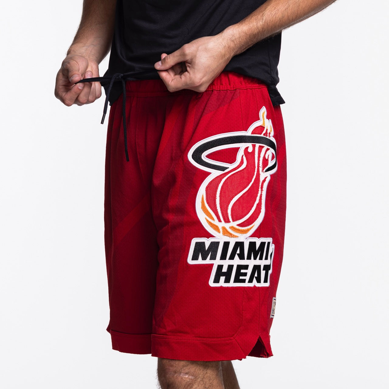 A person adjusts the drawstring on Mitchell & Ness x Purple Brand Miami HEAT Mesh Shorts, made from premium sport mesh with the team's flaming basketball logo. Only the lower torso and upper legs are shown against a white background.