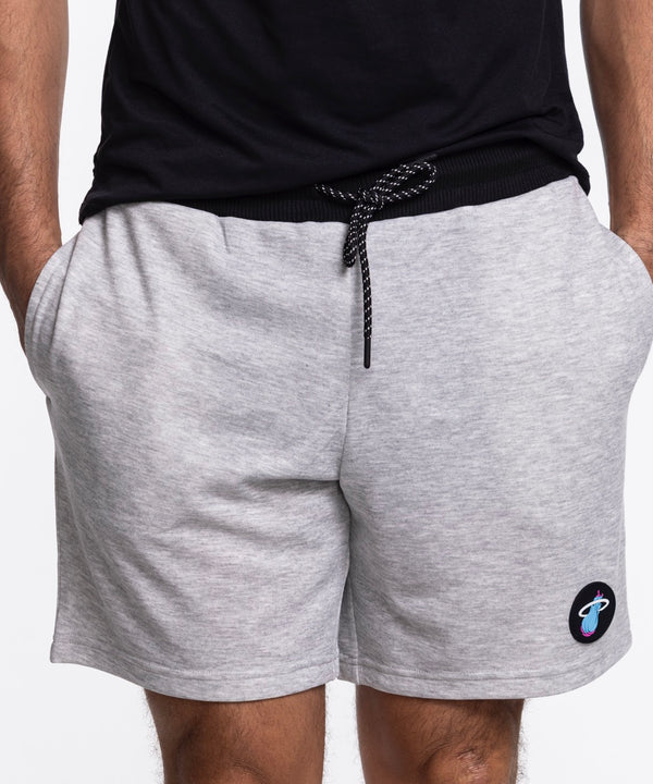 A person wears Concepts Sports Miami HEAT Original Vice Nights Shorts—light gray, relaxed fit with drawstring, hands in pockets—plus a black t-shirt. The polyester cotton shorts have a small round patch of a colorful planet near the hem.