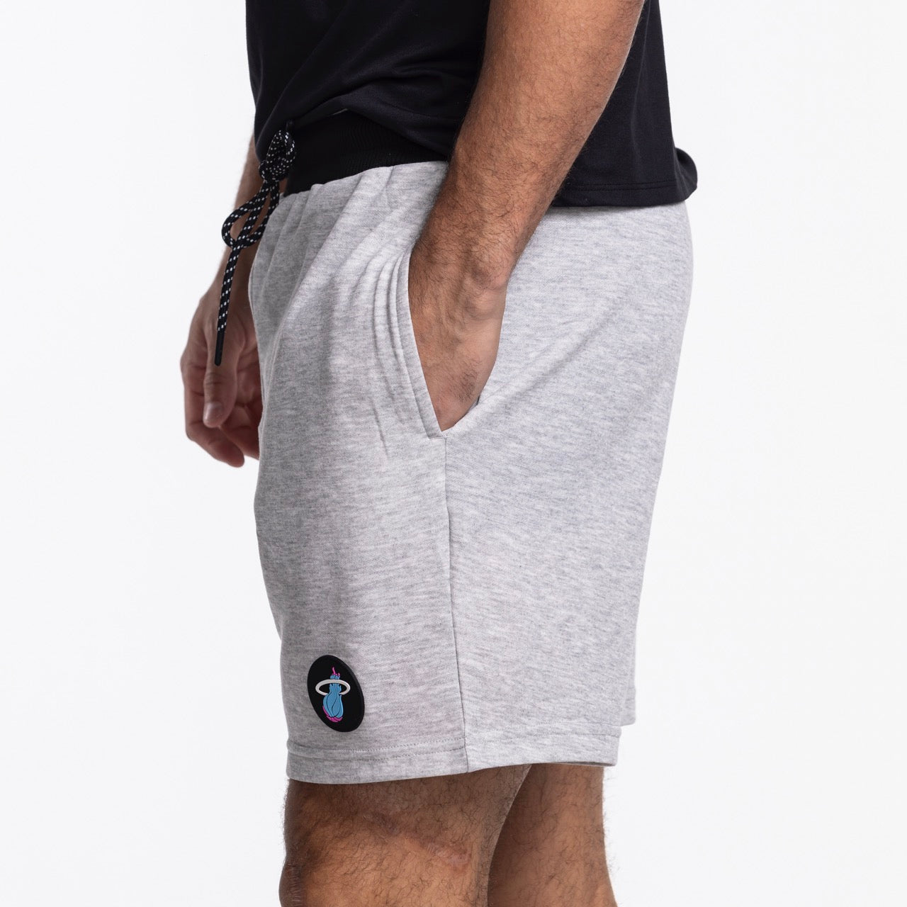 A person wears Concepts Sports Miami HEAT Original Vice Nights Shorts—relaxed fit, light gray polyester-cotton, small colorful logo patch on the left hem—with hands in pockets and a black t-shirt; only lower torso and legs are visible.
