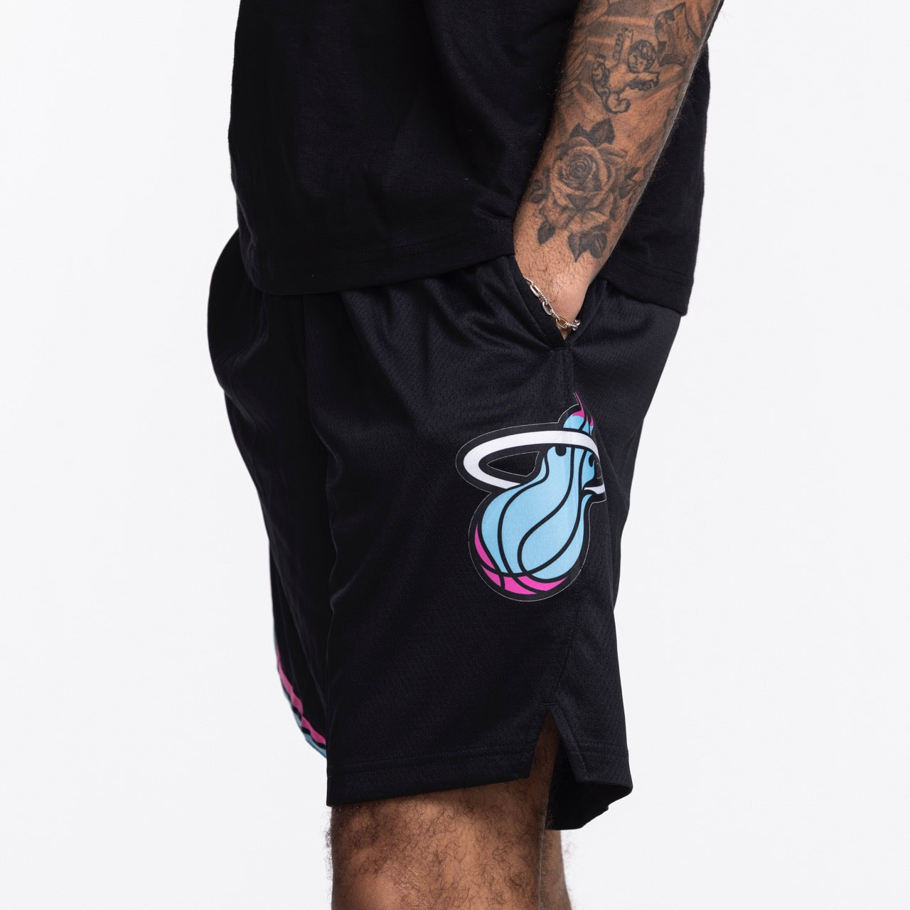 A person with a tattooed arm wears Nike Miami HEAT Original Vice Nights Swingman Shorts in black polyester, featuring a large blue, pink, and white team logo. One hand is in their pocket against a white background. Wash with like colors.