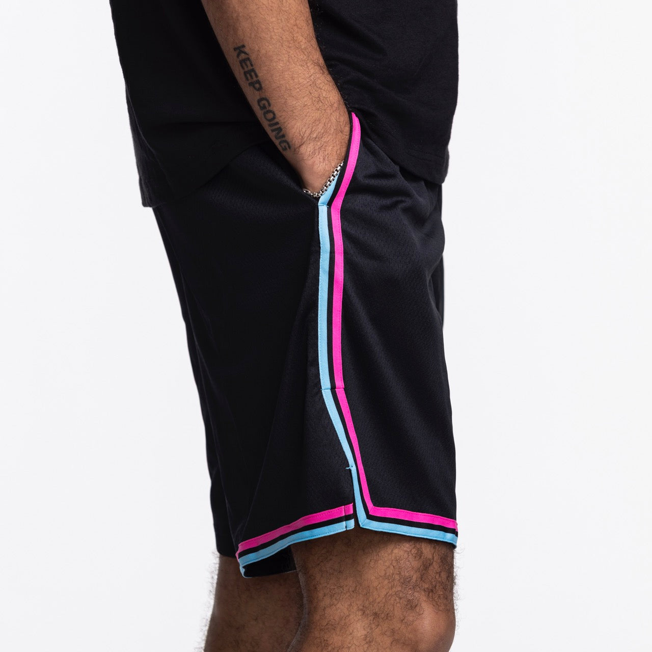 A person wears Nike Miami HEAT Original Vice Nights Swingman Shorts by Nike—standard fit, black with pink and blue trim, paired with a black shirt. Their left hand in pocket reveals a KEEP GOING tattoo. Shorts are polyester; wash with like colors.