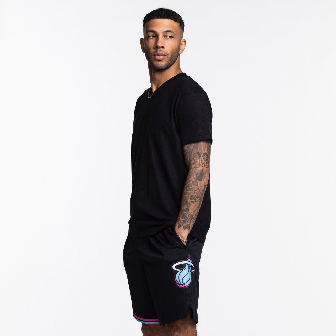 A man with short hair and visible arm tattoos stands sideways in a black t-shirt and Nike Miami HEAT Original Vice Nights Swingman Shorts by Nike, made from polyester, against a plain white background.