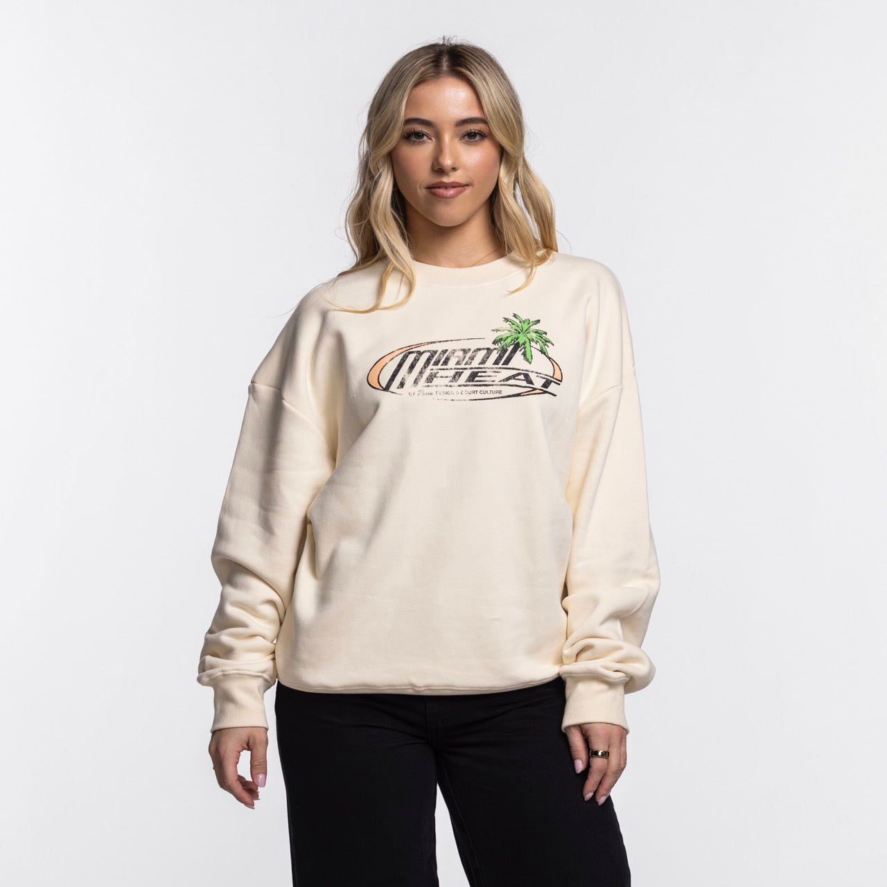 A woman with long blonde hair wears a cream Court Culture x Duvin Sun Crew Sweater and black pants, standing against a plain white background.