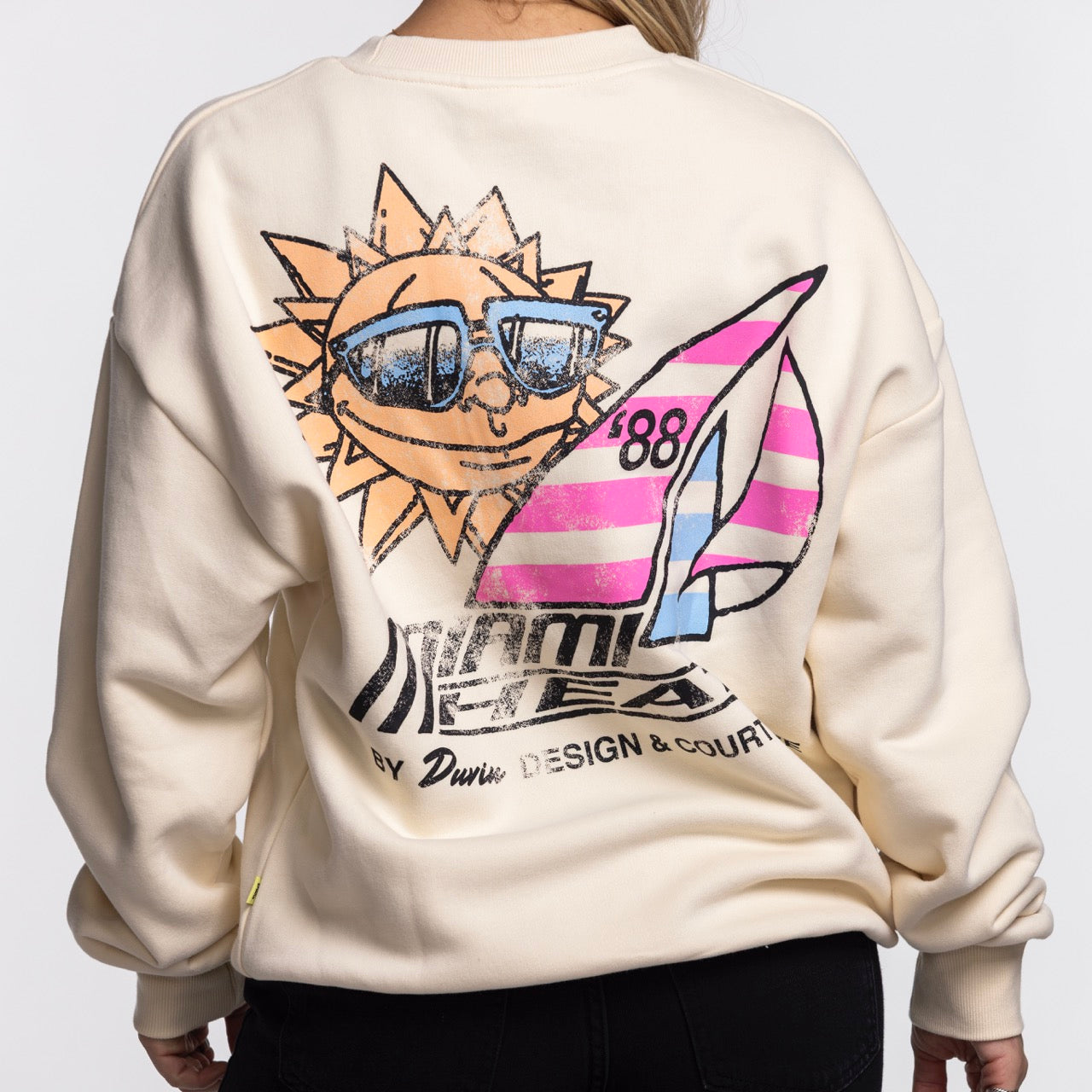 A person wears the Court Culture x Duvin Sun Crew Sweater by Court Culture, a cream sweatshirt with a bold back graphic of a smiling sun, pink-striped sailboat, and retro summer-inspired text.