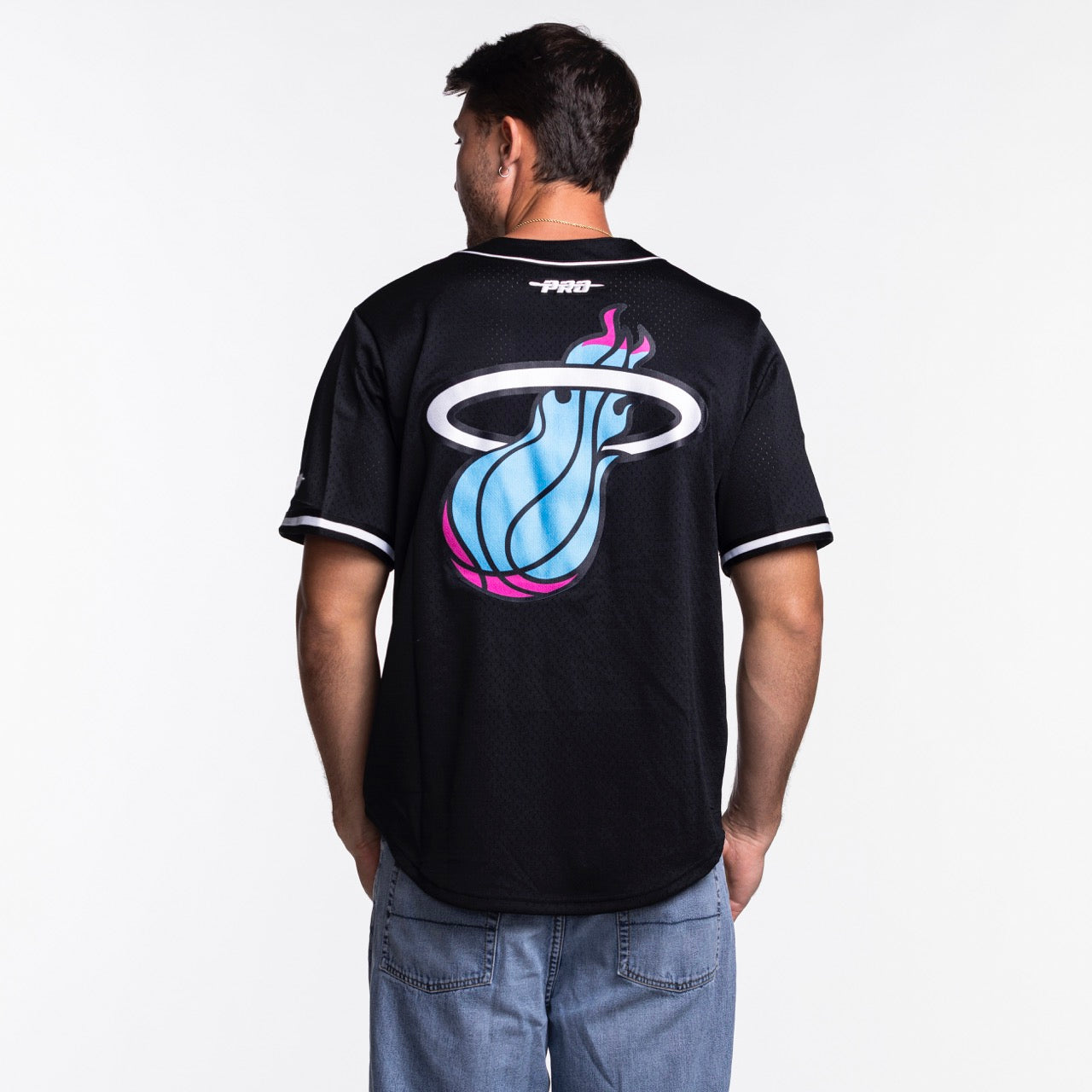 A man faces away, wearing a Pro Standard Miami HEAT Original Vice Nights Baseball Jersey in black polyester with a bold logo on the back. The standard-fit jersey by Pro Standard pairs with light blue jeans, standing out against a plain white background.