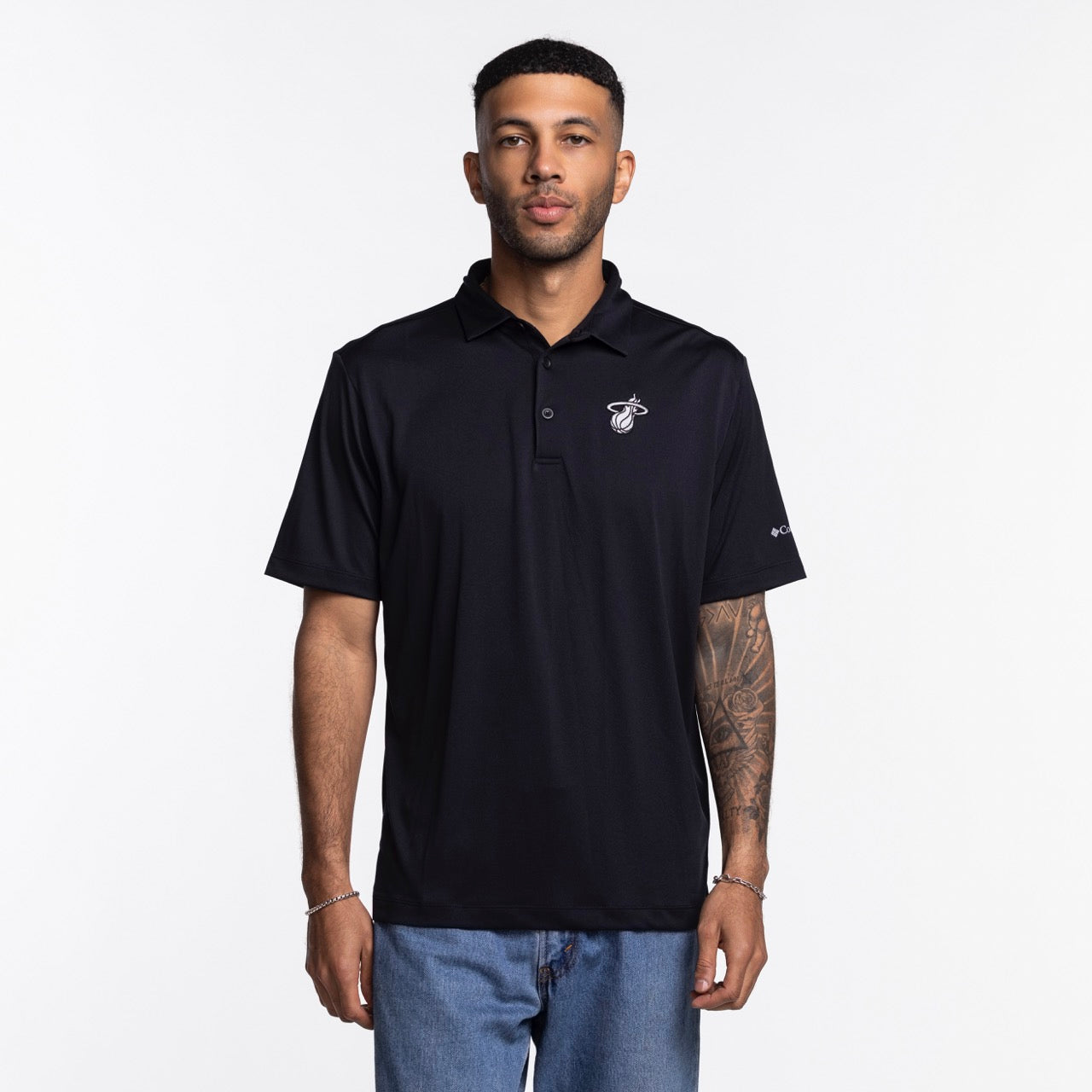 A man with short hair, trimmed beard, and tattooed forearm wears the Columbia Outdoor Miami HEAT Original Vice Nights Tonal Polo—black, standard fit, short-sleeve—paired with light blue jeans against a plain white background.