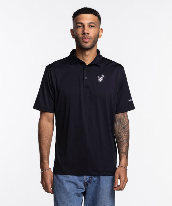 A man with short hair, trimmed beard, and tattooed forearm wears the Columbia Outdoor Miami HEAT Original Vice Nights Tonal Polo—black, standard fit, short-sleeve—paired with light blue jeans against a plain white background.